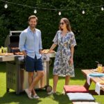 Muž a žena u grilu na zahradě během letního barbecue, obklopeni zelení a světelnými řetězy.