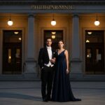 Stylově oblečený pár před historickou filharmonií za soumraku s teplým osvětlením a detailní architekturou