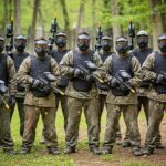 Skupina lidí v paintballovém oblečení s maskami stojí na lesním hřišti připravena k akci.